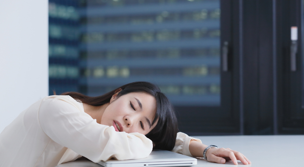 オフィスのデスクで閉じたノートパソコンに突っ伏している女性。【全身(慢性疲労・睡眠負債)】による極度の疲労感や集中力の低下で、限界に達してぐったりしている様子。