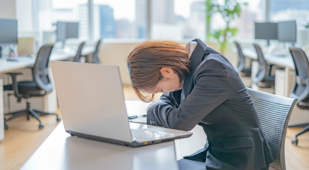 オフィスのデスクで、パソコンの前に突っ伏しているスーツ姿の女性。【心身(極度の過労・自律神経の失調)】により、仕事のストレスや睡眠不足が限界に達し、ぐったりとしている様子。