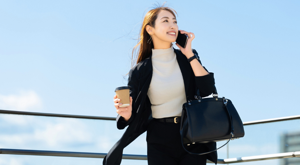 青空の下で女性がコーヒーとバッグを手に持ち、笑顔でスマートフォンで通話しながら颯爽と歩いている画像で、【体幹・下半身(大腰筋・骨盤周辺)】を示し、身体の軸が安定して片側に過度な負担をかけずに荷物を持てるようになり、エネルギッシュに通勤している様子。