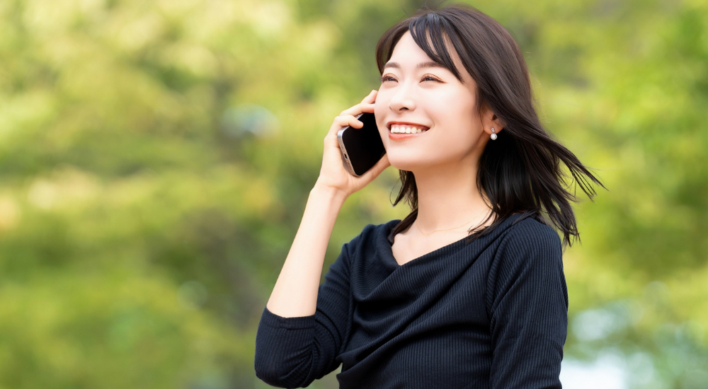 屋外の緑を背景に黒いトップスの女性が笑顔でスマートフォンで通話している画像で、【頸部・顔面(胸鎖乳突筋・表情筋周辺)】を示し、首周りの緊張や自律神経の乱れが整うことで自然と表情も明るくなり、ストレスフリーで健やかな日常生活を送っている様子。
